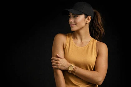 Sportos nő fekete baseball sapkában és matt arany árnyalatú trikóban, arany színű karórával és karkötővel, fekete háttér előtt – prémium fitness lifestyle megjelenés.